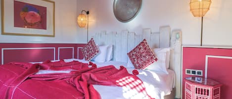 Una cama con una manta roja, almohadas blancas y un cabecero de madera. Hay una mesita de noche roja pequeña con una lámpara y un aplique de pared.