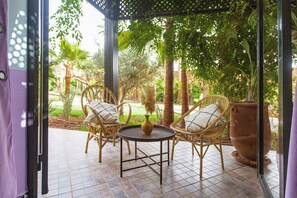 An outdoor patio with wicker furniture and a round table, surrounded by a garden with palm trees and a large pot.