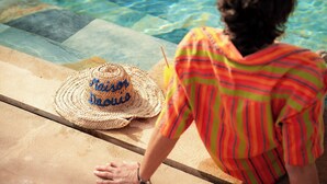 Una persona seduta a bordo piscina, con un cappello di paglia e un drink.