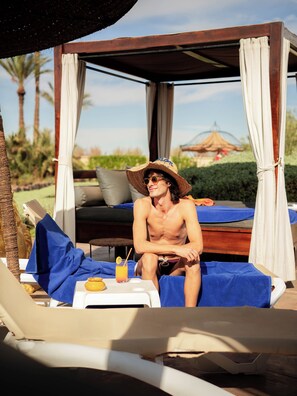 A man sitting on a sunbed in a covered outdoor area with a view of a garden and a building in the background. - Maison Daouia (Marrakech)