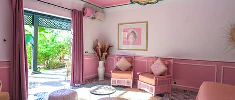 A pink-themed room with a large window, a white pouf, a pink sofa, and a decorative hanging light.