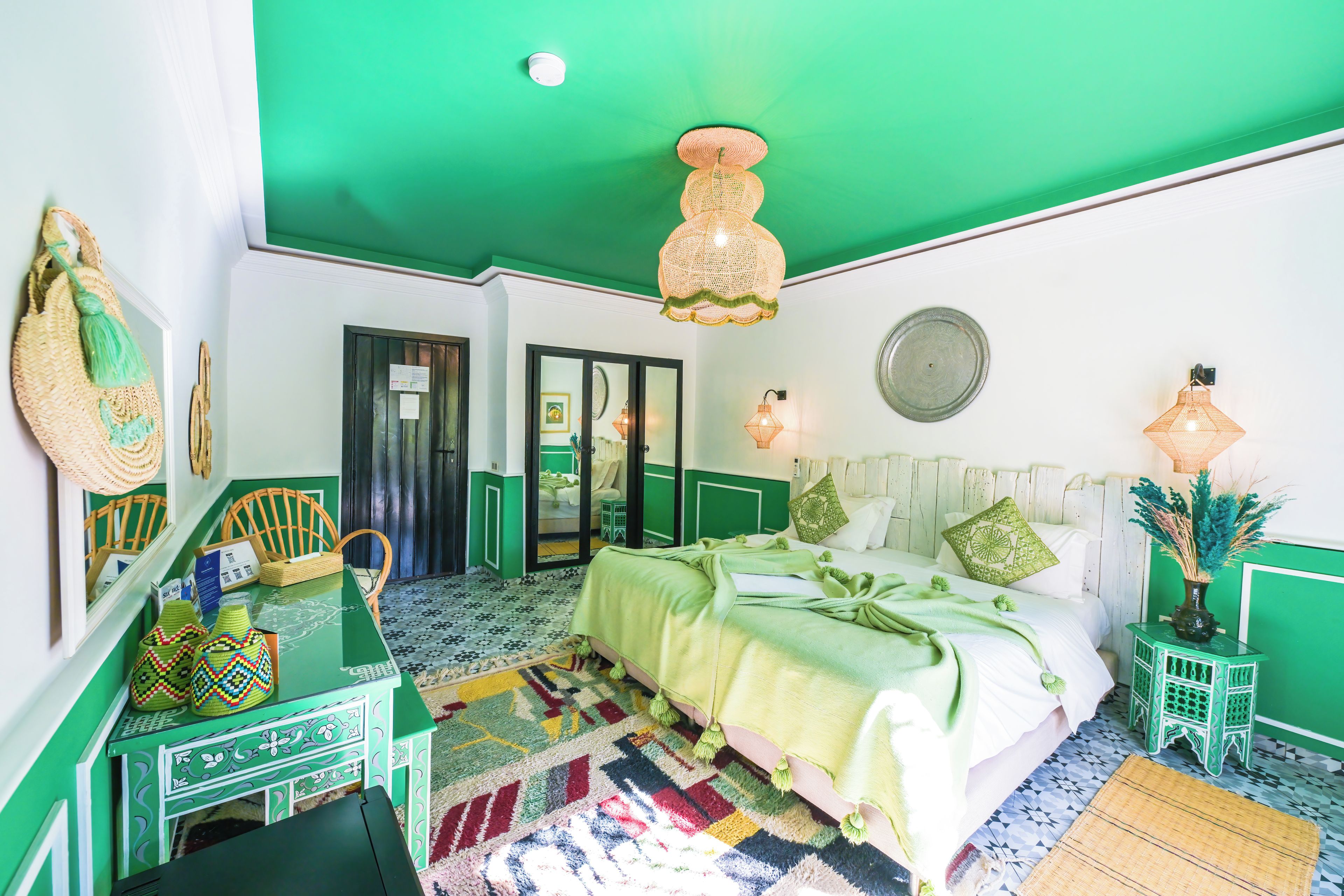 A room with a green ceiling, a bed with green bedding, a small table with chairs, and a mirror on the wall.