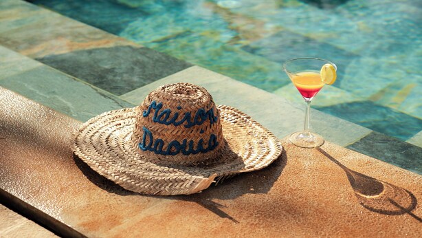 A straw hat with "Maison Daou" embroidered on it, a cocktail glass with a lemon slice, and a poolside setting. - Maison Daouia (Marrakech)