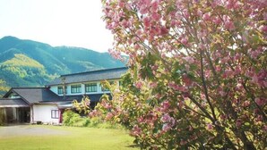 A traditional building with a sloped roof, surrounded by lush greenery and mountains in the background. - MIZUKAMISO (Mizukami)