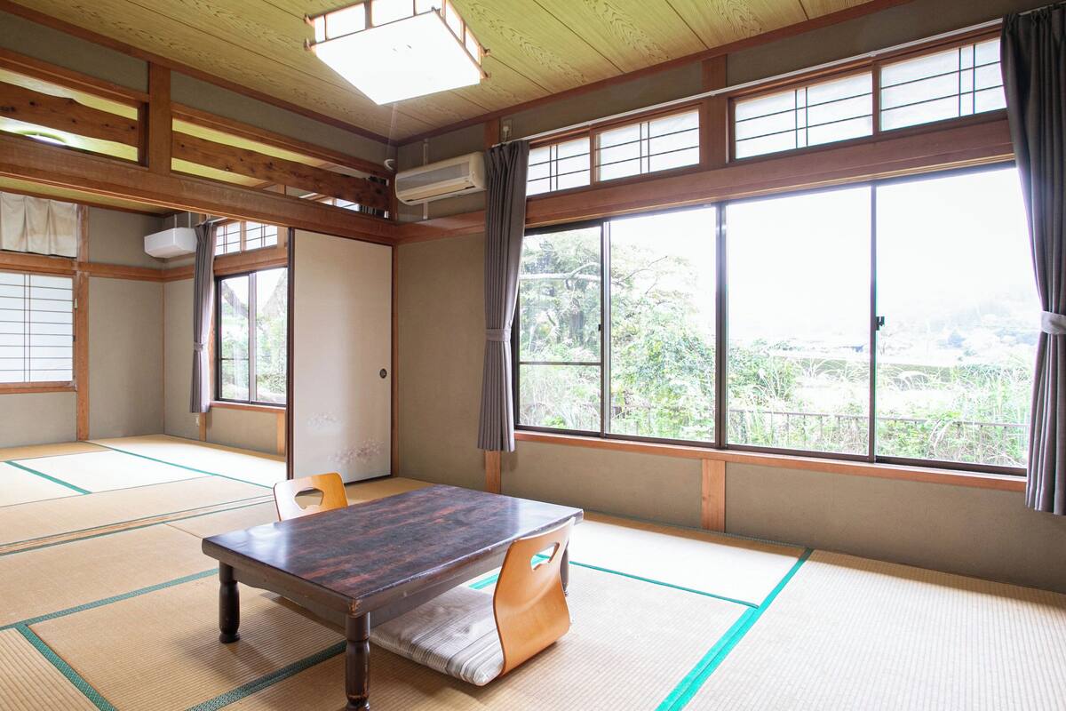 Ein traditioneller japanischer Raum mit Tatami-Böden, einem Holztisch und großen Fenstern mit Vorhängen.
