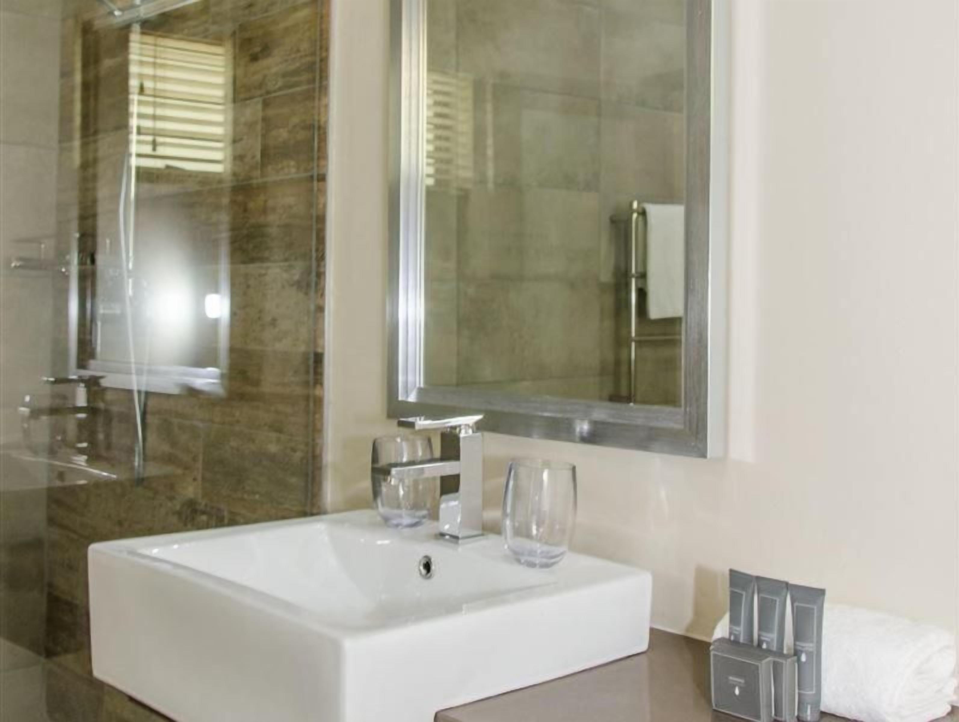 Un baño moderno con un lavamanos grande de color blanco, dos vasos de vidrio, un espejo y un toallero con artículos de aseo.