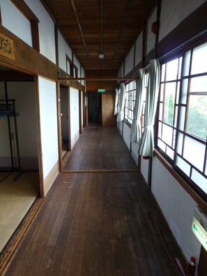 Un couloir doté d’un plancher en bois, de grandes fenêtres avec des rideaux et de portes coulissantes traditionnelles japonaises.