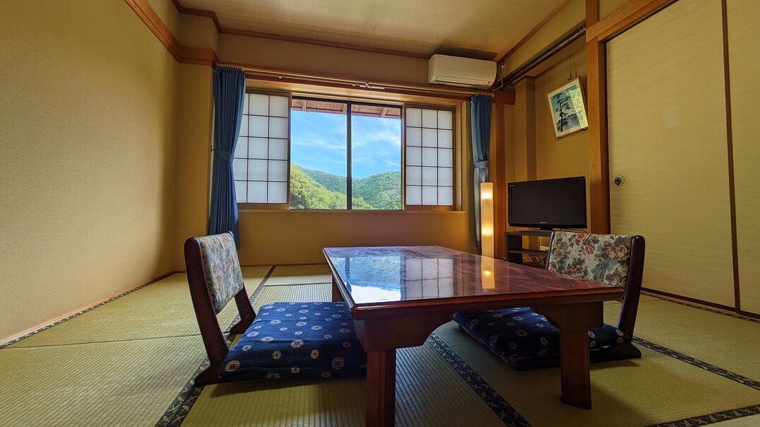 Une pièce de style japonais traditionnel, avec un sol recouvert de tatamis, une table basse, deux chaises, un téléviseur et une fenêtre donnant sur une vue sur la montagne.