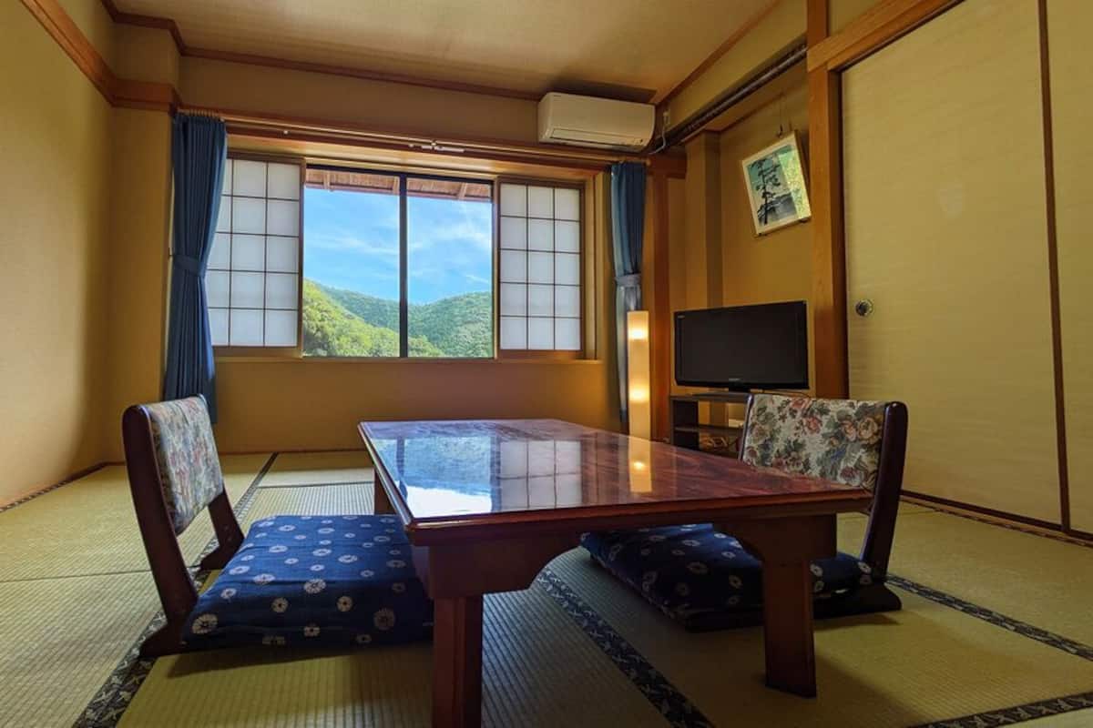 Une pièce de style japonais traditionnel, avec un sol recouvert de tatamis, une table basse, deux chaises, un téléviseur et une fenêtre donnant sur une vue sur la montagne.