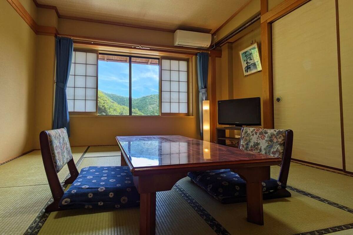 Une pièce de style japonais traditionnel, avec un sol recouvert de tatamis, une table basse, deux chaises, un téléviseur et une fenêtre donnant sur une vue sur la montagne.