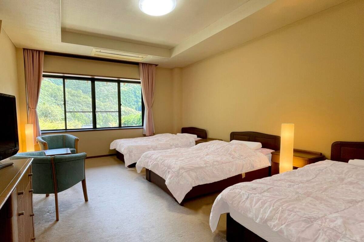 Une chambre d’hôtel avec trois lits, un bureau avec une télévision, une chaise et une fenêtre avec des rideaux.