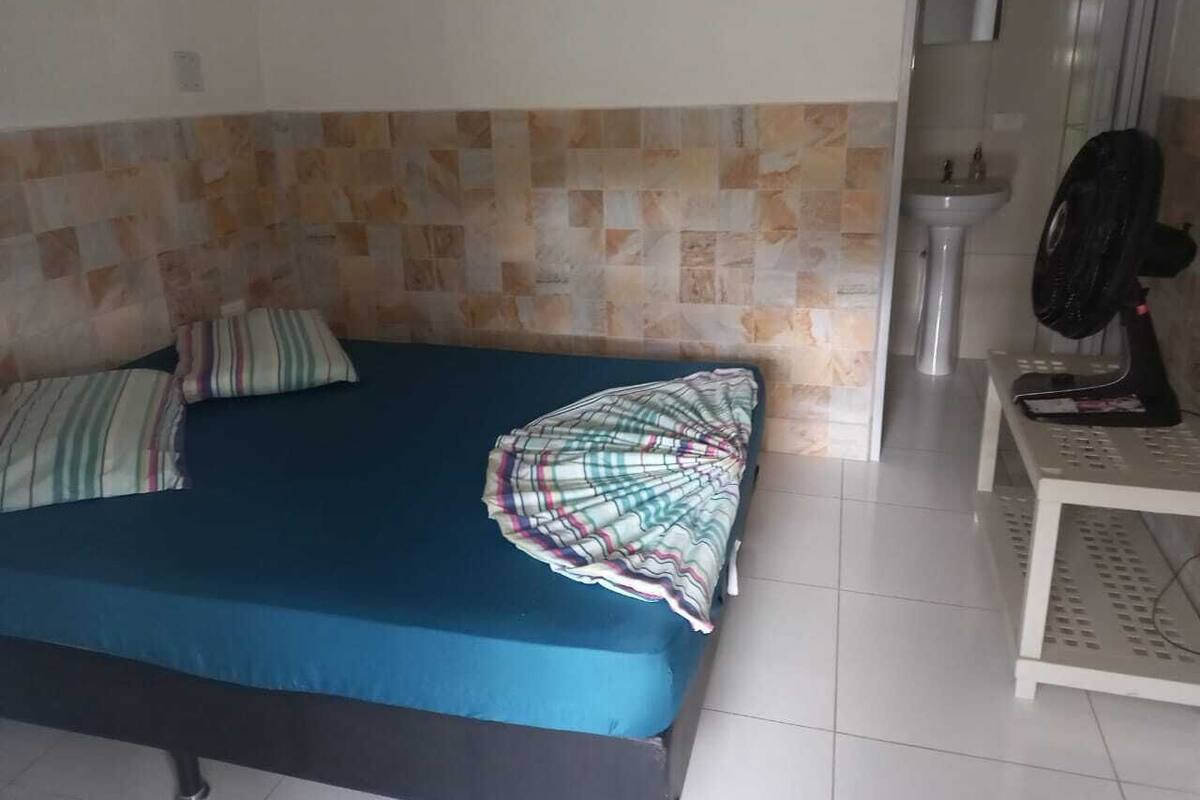 Een slaapkamer met een bed, kussens, een nachtkastje en een ventilator.
