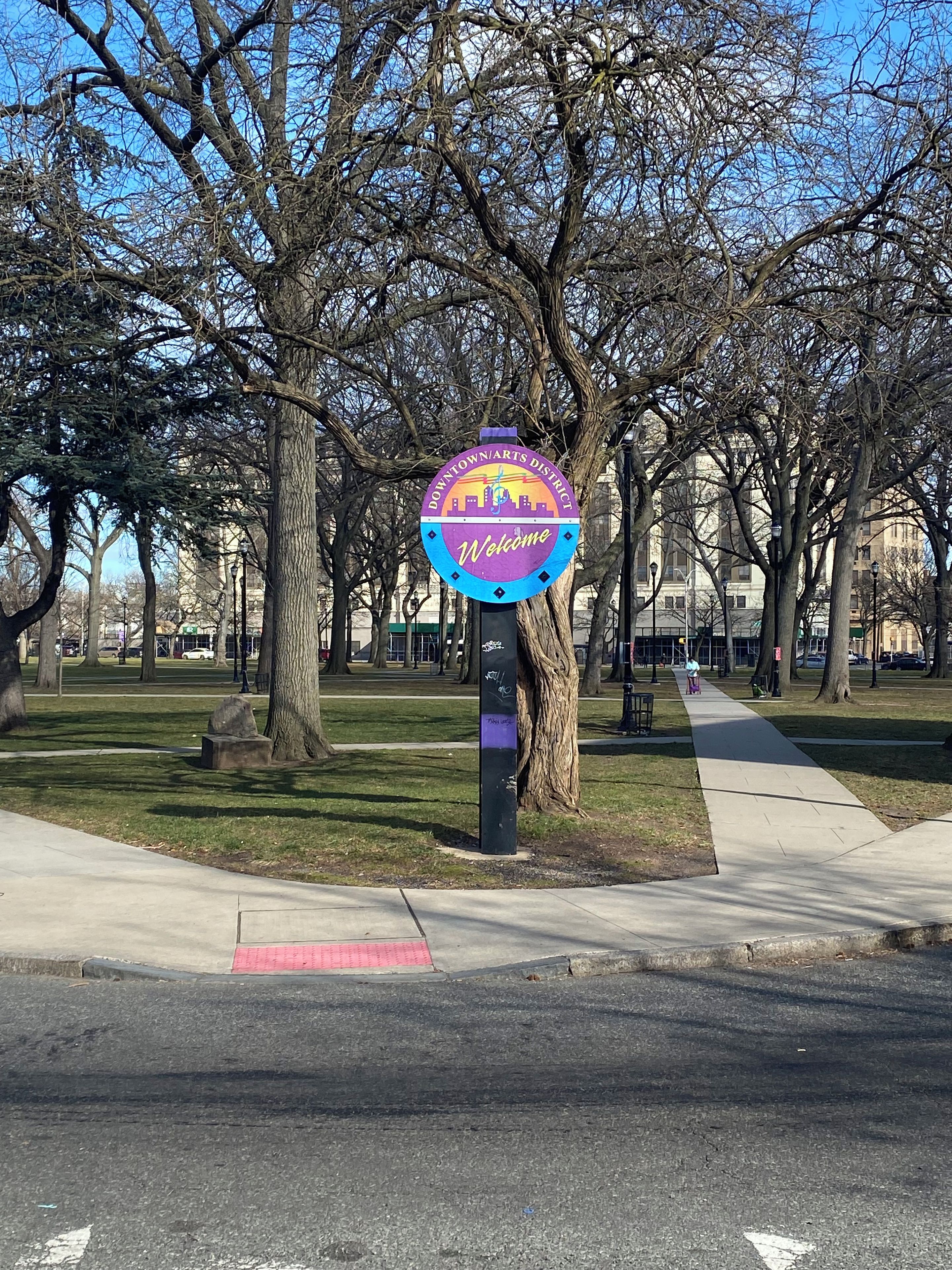 A welcome sign for Downtown Arts District with a clear path and trees in the background.