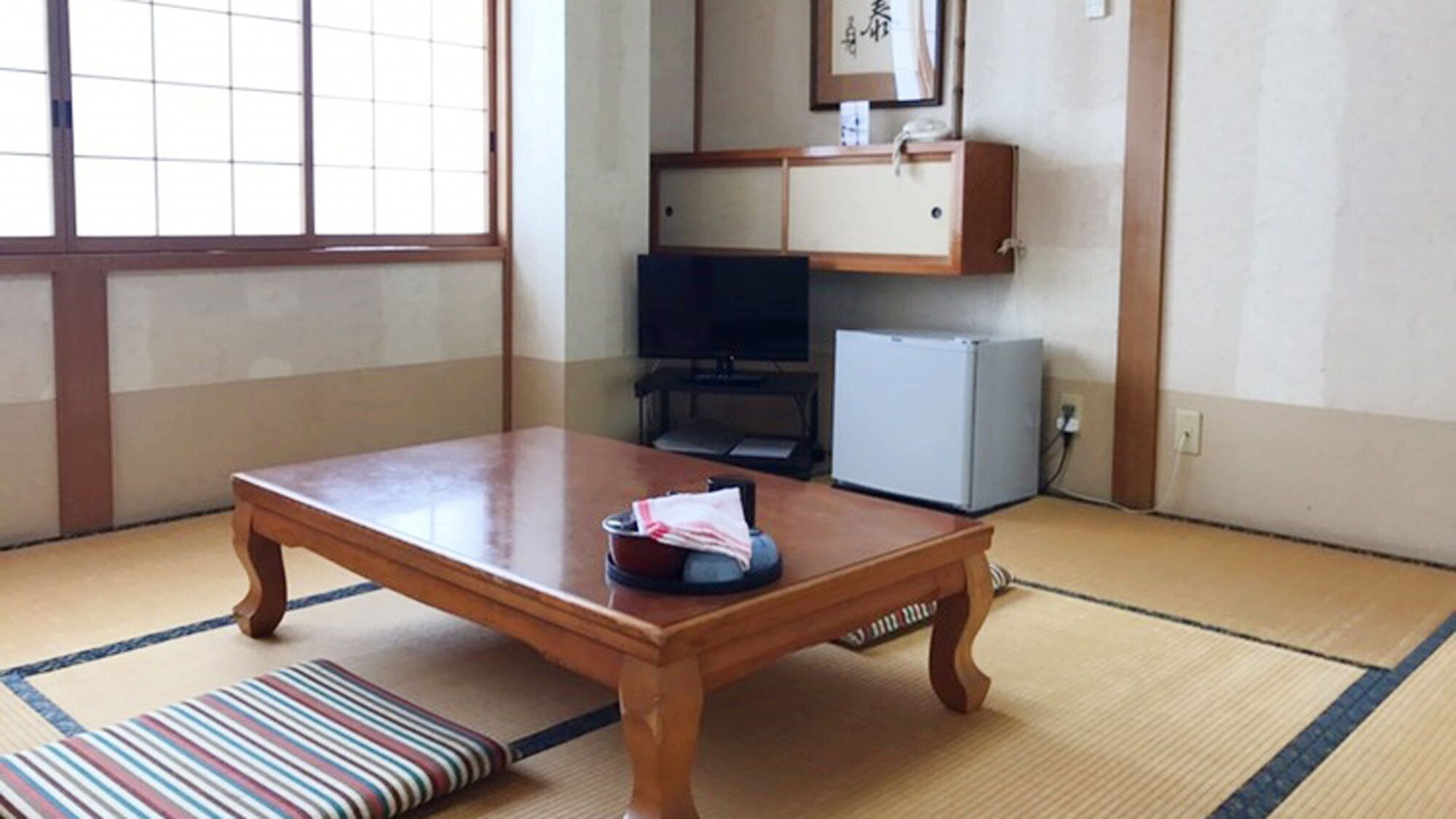 Ein traditioneller japanischer Raum mit Tatami-Boden, einem Holztisch, einem Fernseher und einem kleinen Kühlschrank.