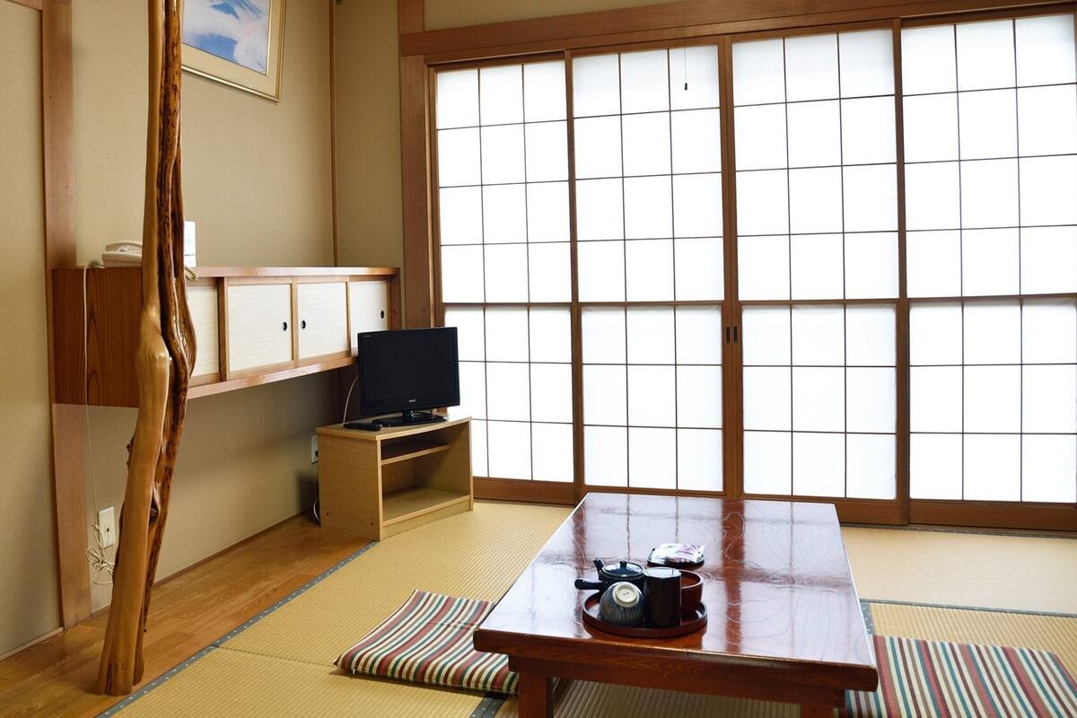 Una tradizionale stanza giapponese con pavimento in tatami, un televisore su un mobile basso, un tavolo di legno con un vassoio e una porta scorrevole shoji.