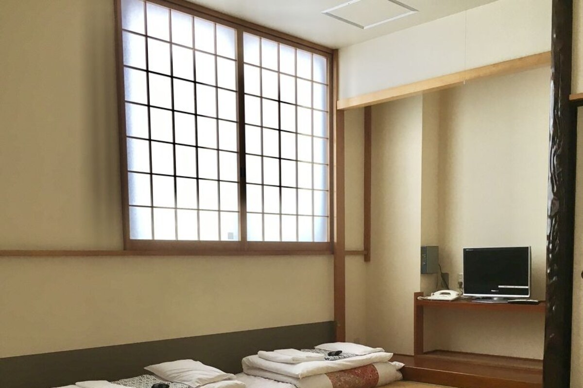 A traditional Japanese room with tatami mats, a futon bed, a television on a wooden stand, and a sliding door.
