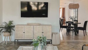 En moderne stue med et stort fladskærms-tv monteret på væggen, et rattan-konsolbord med dekorative planter, et marmorbord og en gulvtæppe med mønster.