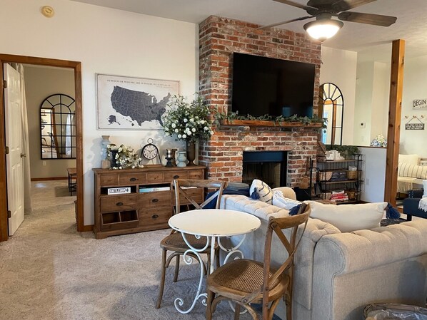 A cozy living room with a brick fireplace, a flat-screen TV mounted above it, and a dining area with a round table and wicker chairs.