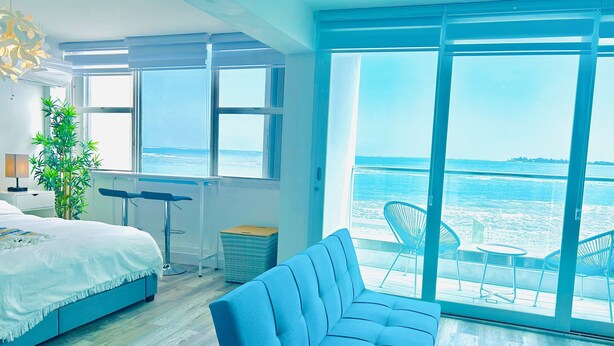 Un dormitorio moderno con una cama grande, un sofá azul, una mesa de comedor y vistas al mar a través de amplias ventanas.