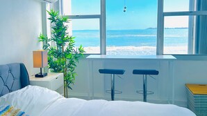Un dormitorio con un amplio ventanal con vistas al mar, una cama, un escritorio con lámpara y una planta en maceta.