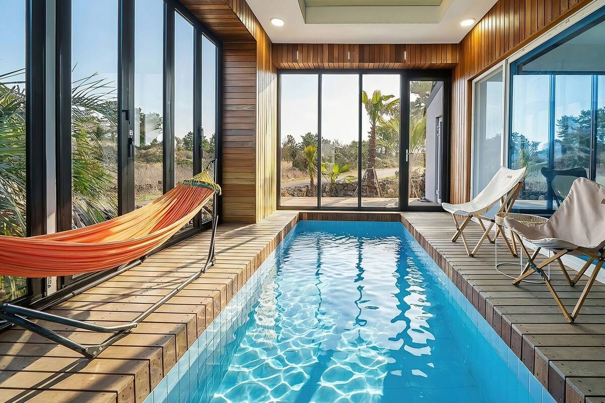 A modern indoor pool area with a hammock, lounge chairs, and large windows offering a view of the outdoors.