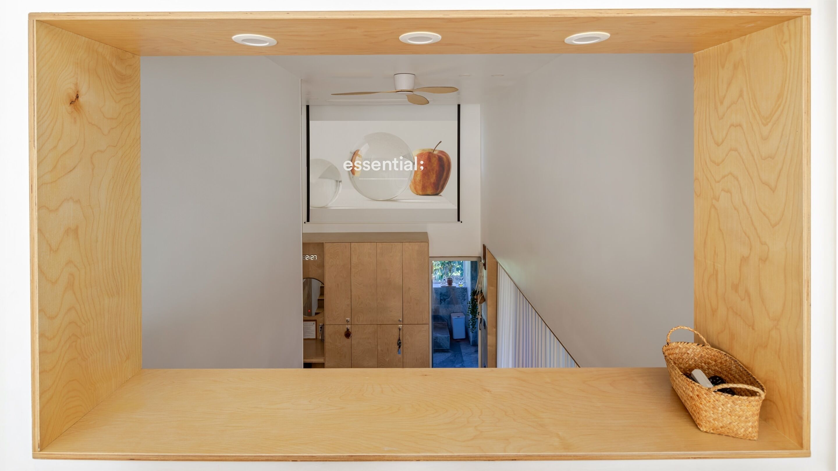 Una habitación minimalista con suelo de madera, ventilador de techo, lámpara de pared y una cesta tejida en el lado derecho.