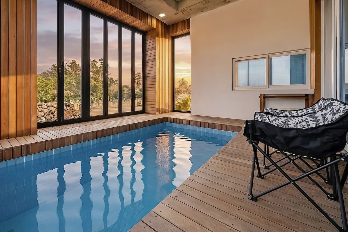 A modern indoor swimming pool with wooden decking, large windows offering a view of the outdoors, and a folding chair by the pool.