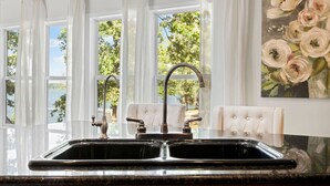 A kitchen with a double sink, white curtains, and a view of trees and water outside.