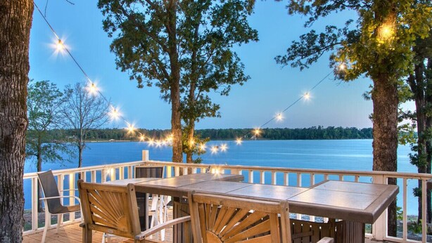 A wooden deck with a table and chairs overlooking a lake, string lights, and trees.