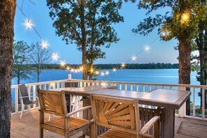 A wooden deck with a table and chairs overlooking a lake, string lights, and trees.