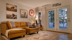 A living room with a brown leather sofa, a patterned rug, and a guitar. There are framed pictures on the wall and a sign that reads "Heleads to Still Waters".