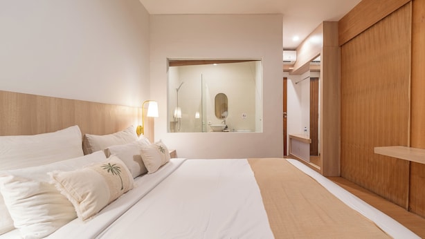 Une chambre d’hôtel moderne avec un grand lit, une armoire en bois, un miroir et une salle de bain visible à travers une porte ouverte.