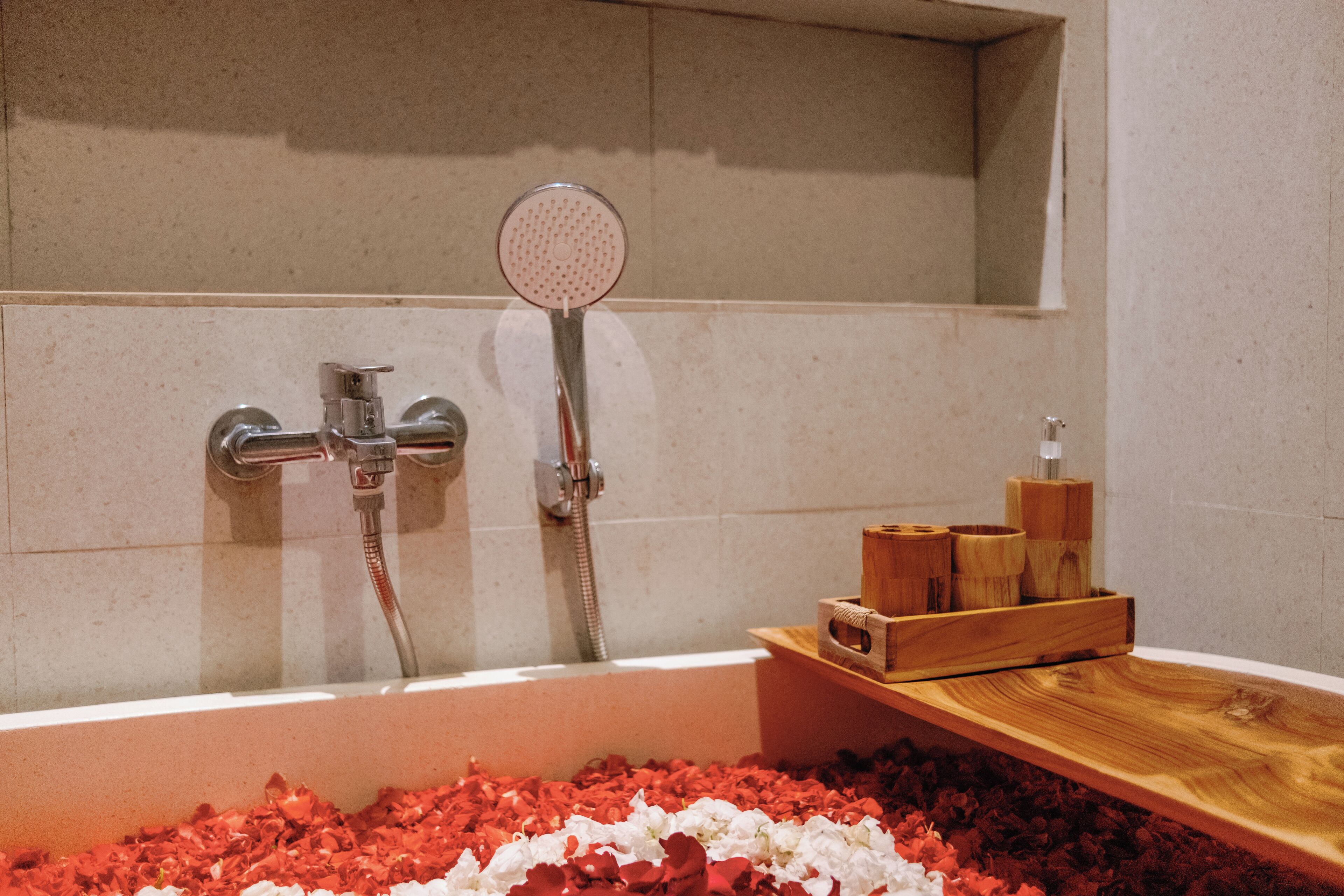 Una bañera llena de pétalos de rosa, un cabezal de ducha y contenedores de madera sobre un estante de madera.