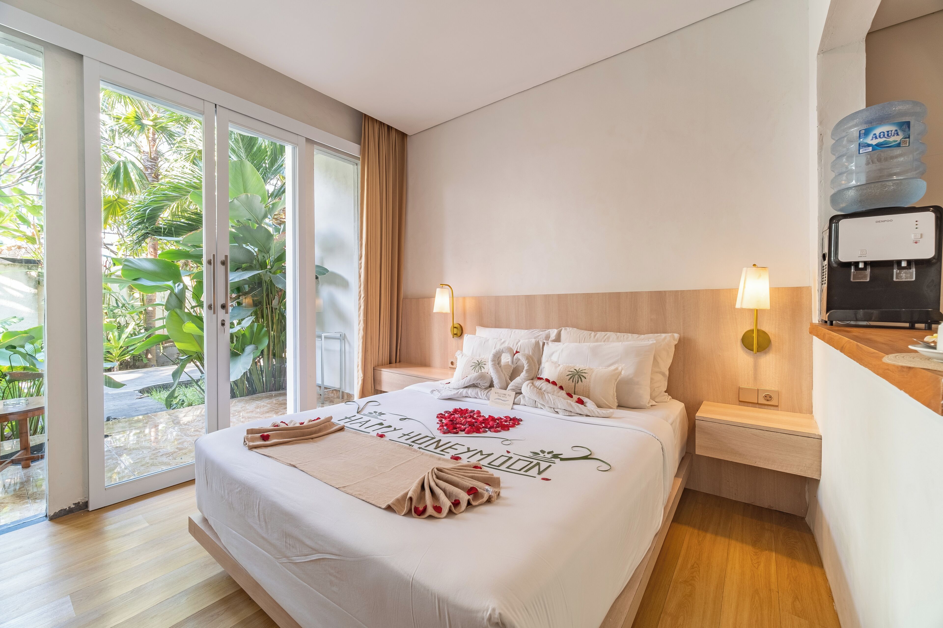 A hotel room with a bed, two bedside tables, a water dispenser, and a view of greenery through the sliding glass door.