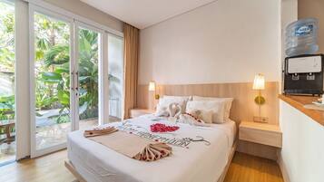 A hotel room with a bed, two bedside tables, a water dispenser, and a view of greenery through the sliding glass door.