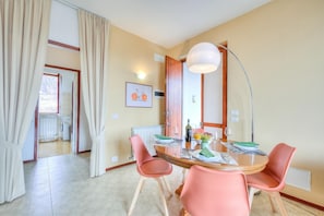 A dining area with a round table, pink chairs, a lamp, and a view of a kitchen and bathroom through an open door.