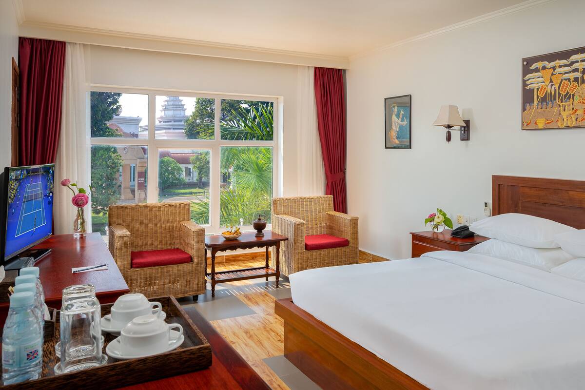 Een hotelkamer met een groot bed, twee rieten stoelen, een klein tafeltje met een vaas, een televisie en een schilderij aan de muur.