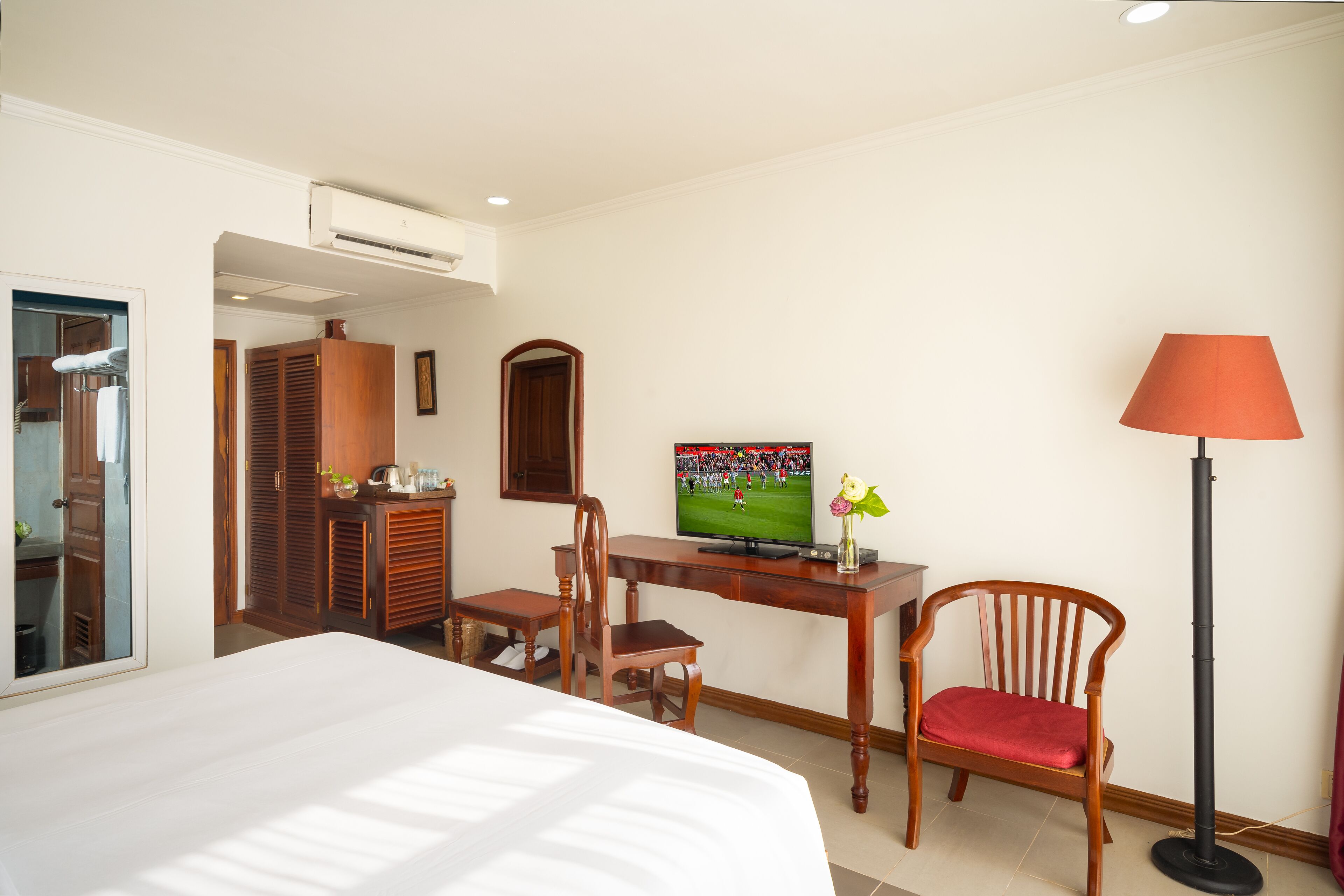 A hotel room with a bed, a desk with a television, a chair, a lamp, and a wardrobe.