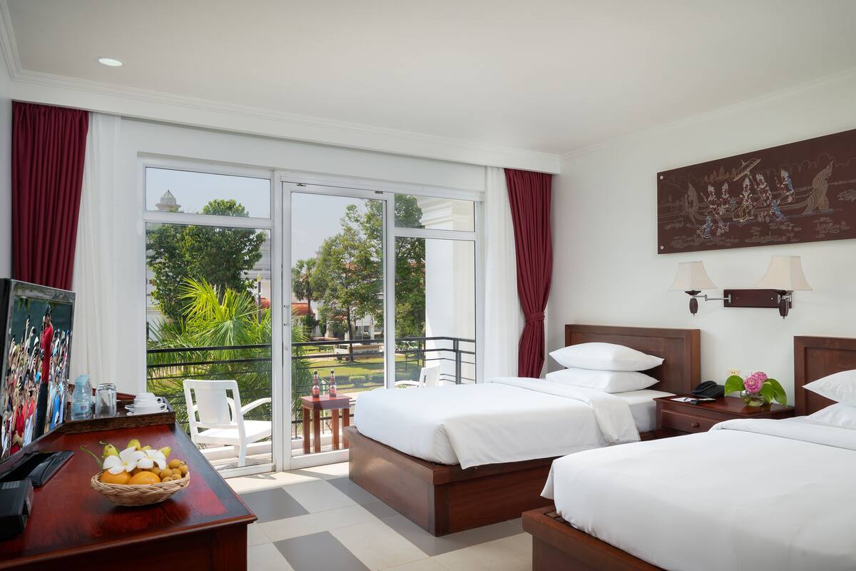 Een hotelkamer met twee bedden, een balkon met uitzicht op groen, een bureau met een mand fruit en een schilderij aan de muur.