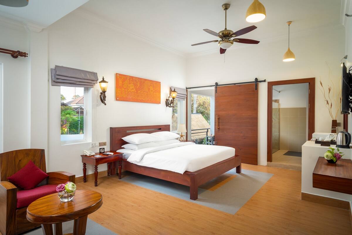 Hotelkamer met een houten bed, een bureau, een stoel, een klein tafeltje met een vaas, een plafondventilator en uitzicht op buiten.