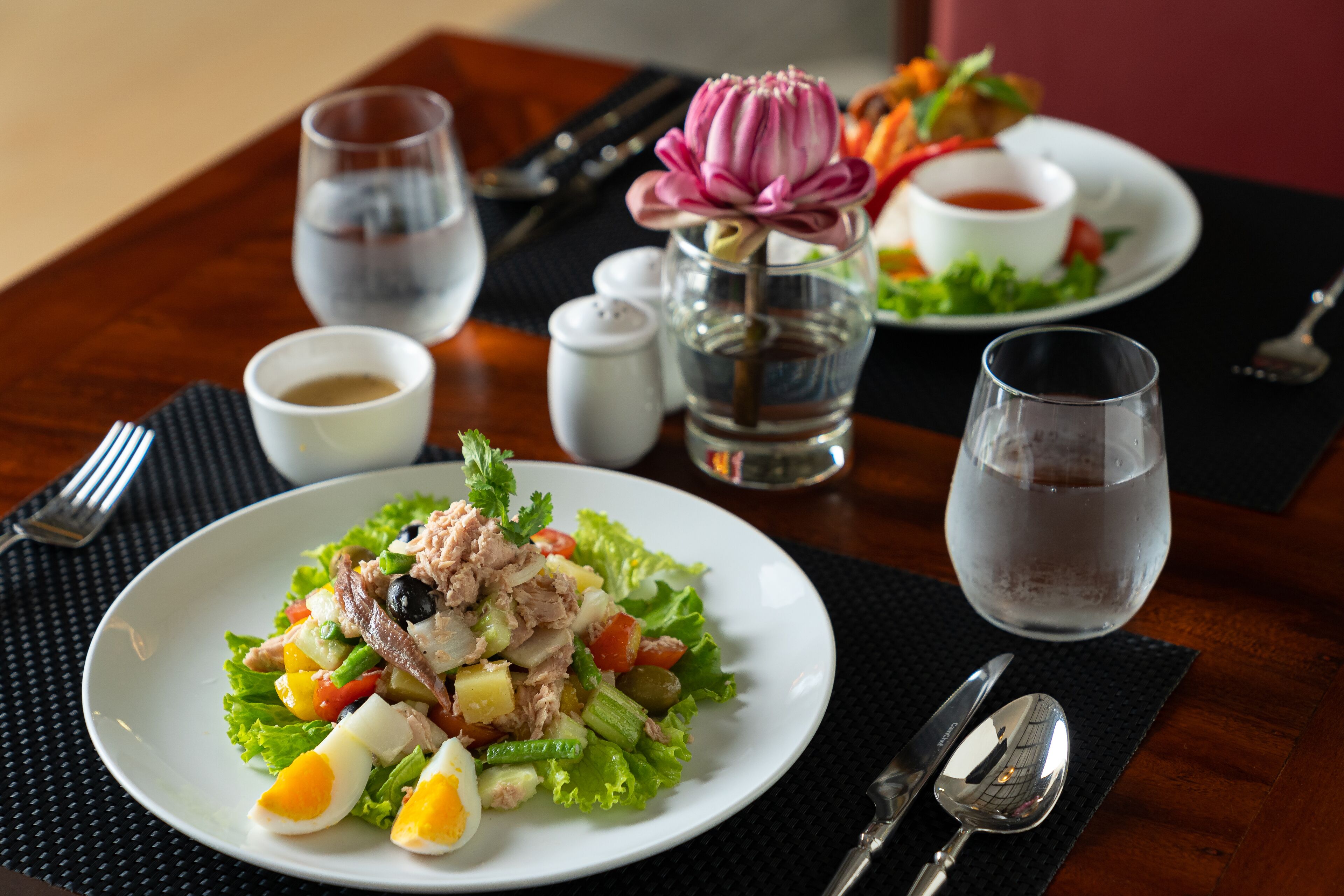 A dining table with a salad, a glass of water, a cup of coffee, and a flower vase.