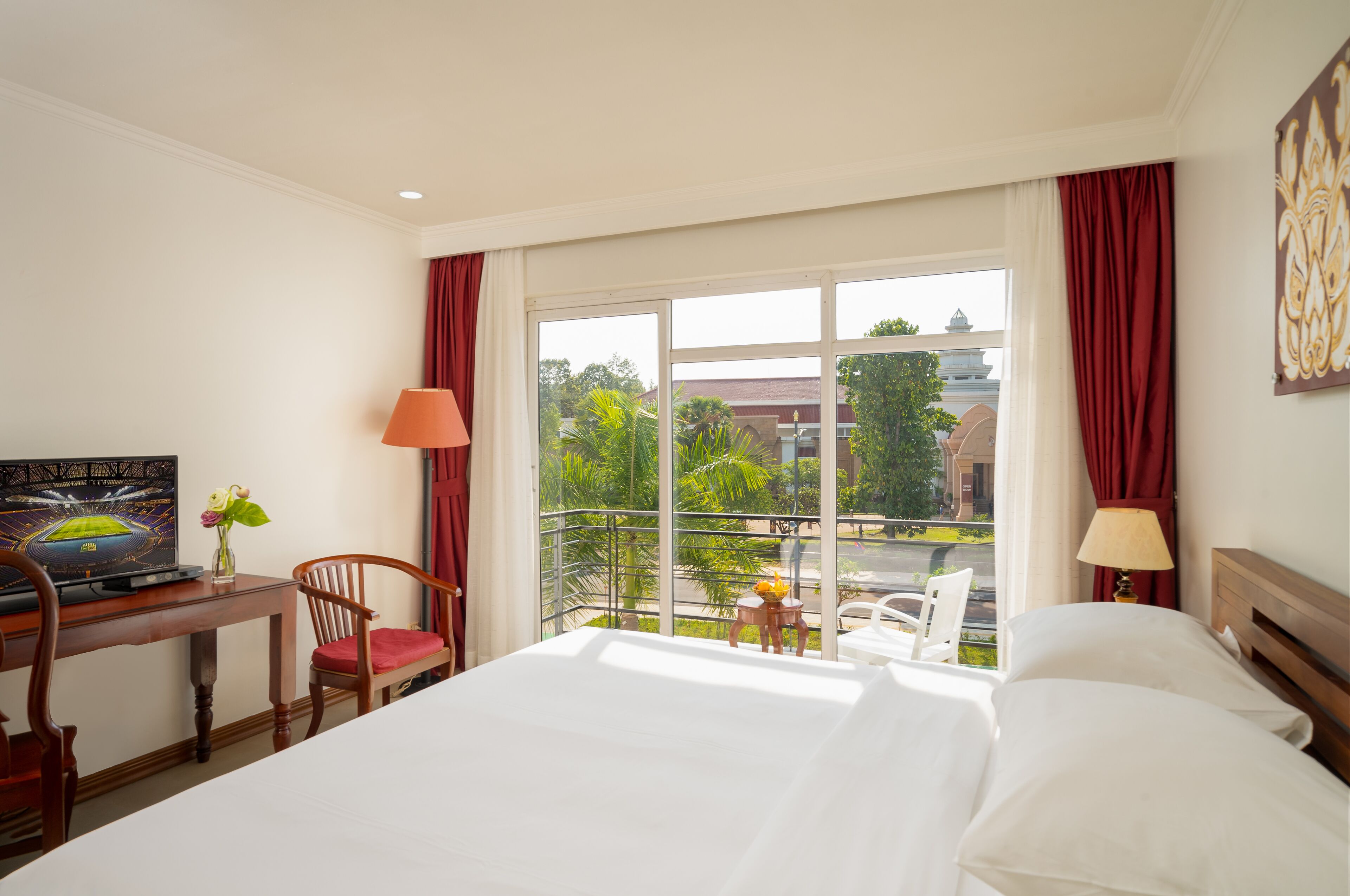 A hotel room with a bed, a desk with a laptop, a chair, a small table with a vase, and a balcony with a view of greenery and buildings.