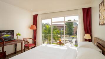 A hotel room with a bed, a desk with a laptop, a chair, a small table with a vase, and a balcony with a view of greenery and buildings.