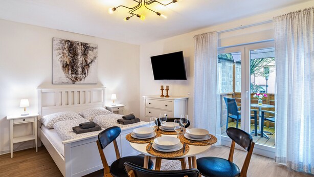 Habitación de hotel con cama, mesita de noche, televisor, mesa de comedor con sillas y balcón con vistas.