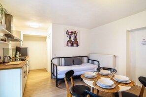 A compact room with a kitchenette, a dining table set for two, a sofa, and a painting on the wall.