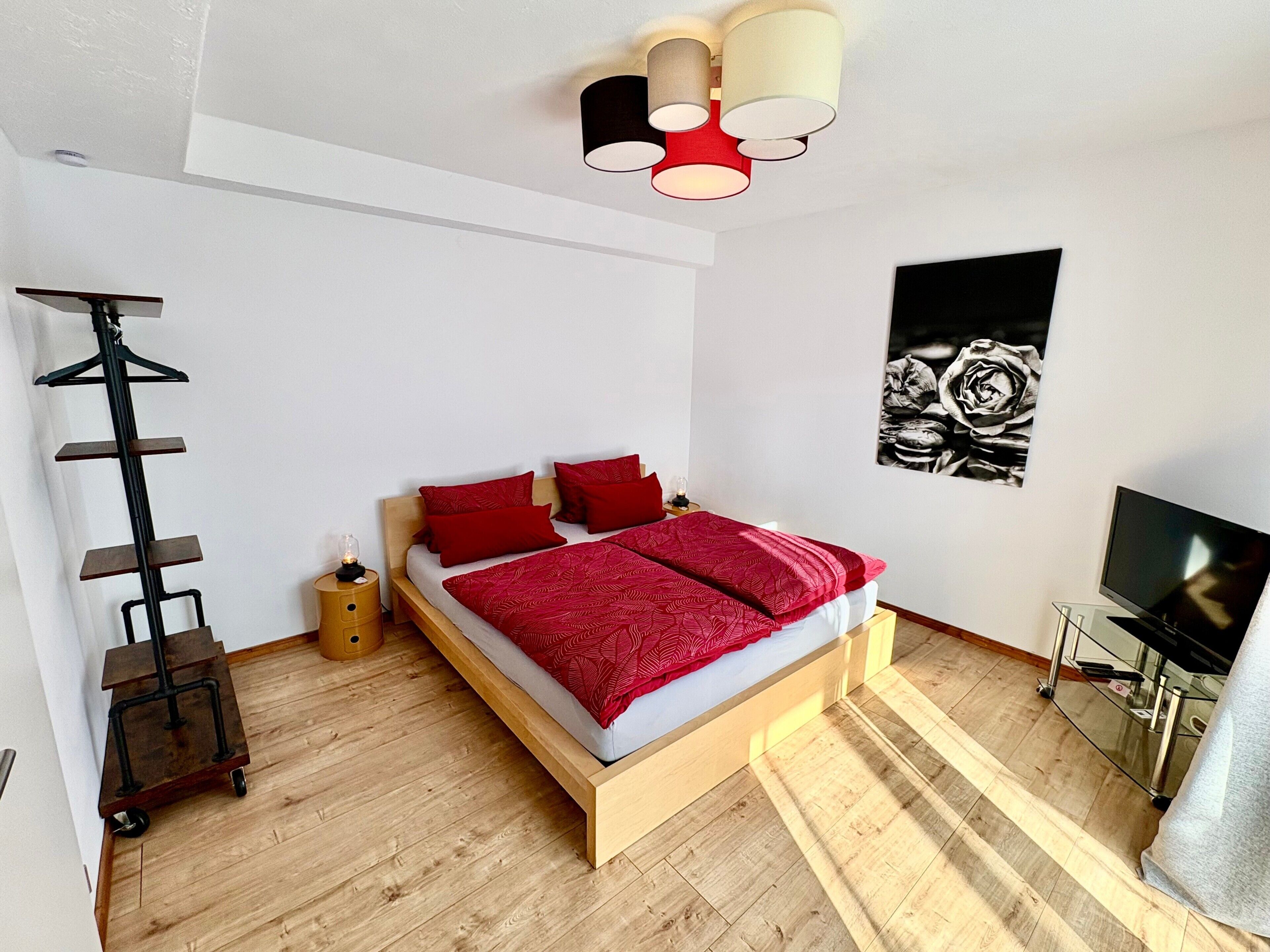 Een moderne slaapkamer met een houten bed, rode beddengoed, een zwarte tv-meubel en een plafondlamp.