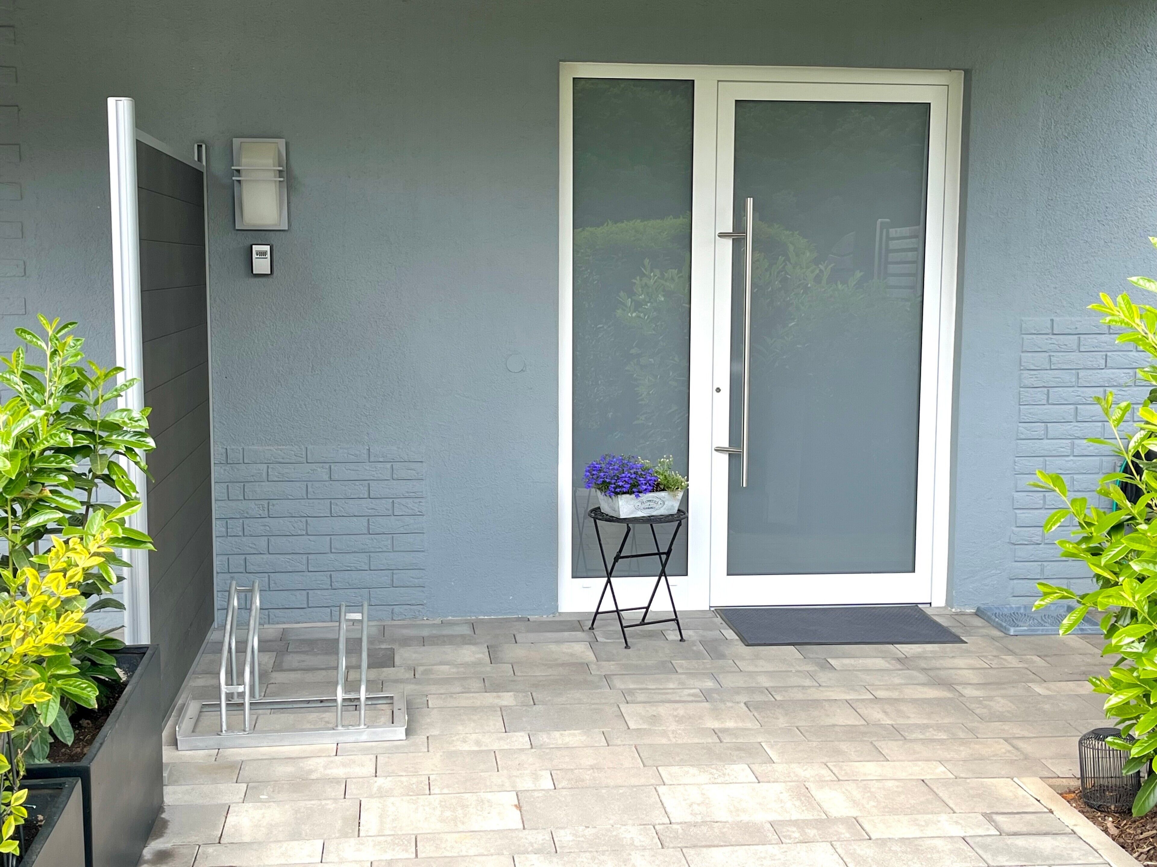 A modern exterior with a grey brick wall, a glass door with a white frame, a small table with a potted plant, and a metal ladder.