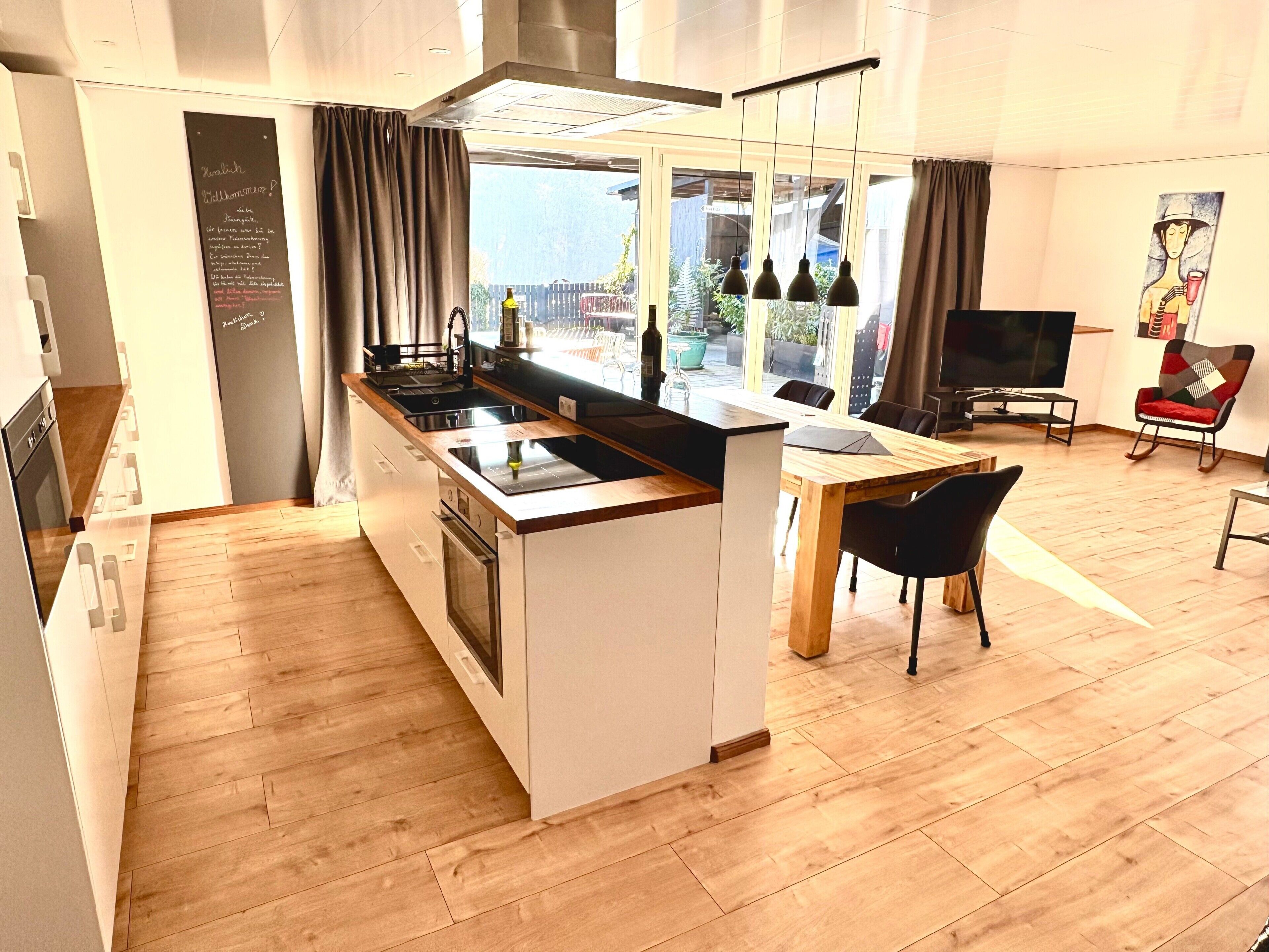 Eine moderne Küche mit einer zentralen Kochinsel, ein Essbereich mit Holztisch und Stühlen sowie ein Wohnbereich mit Fernseher und Kunstwerken.