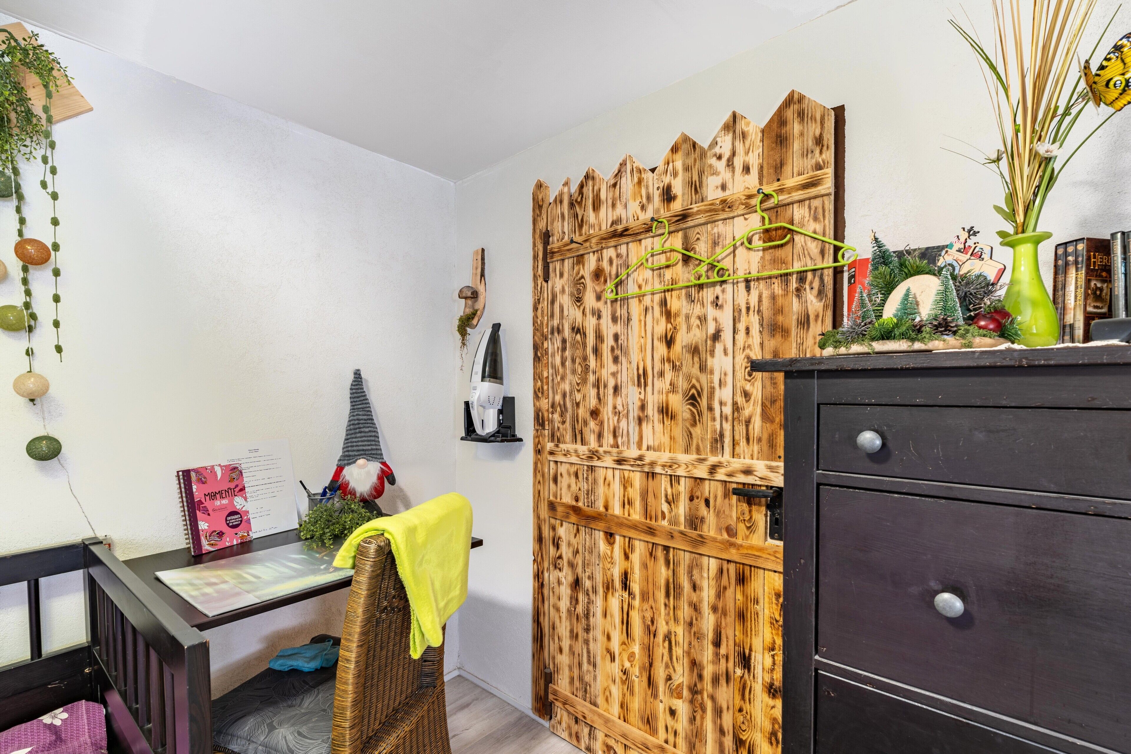 Ein Zimmer mit Holztür, Schreibtisch mit Stuhl, kleines Regal und Kommode.