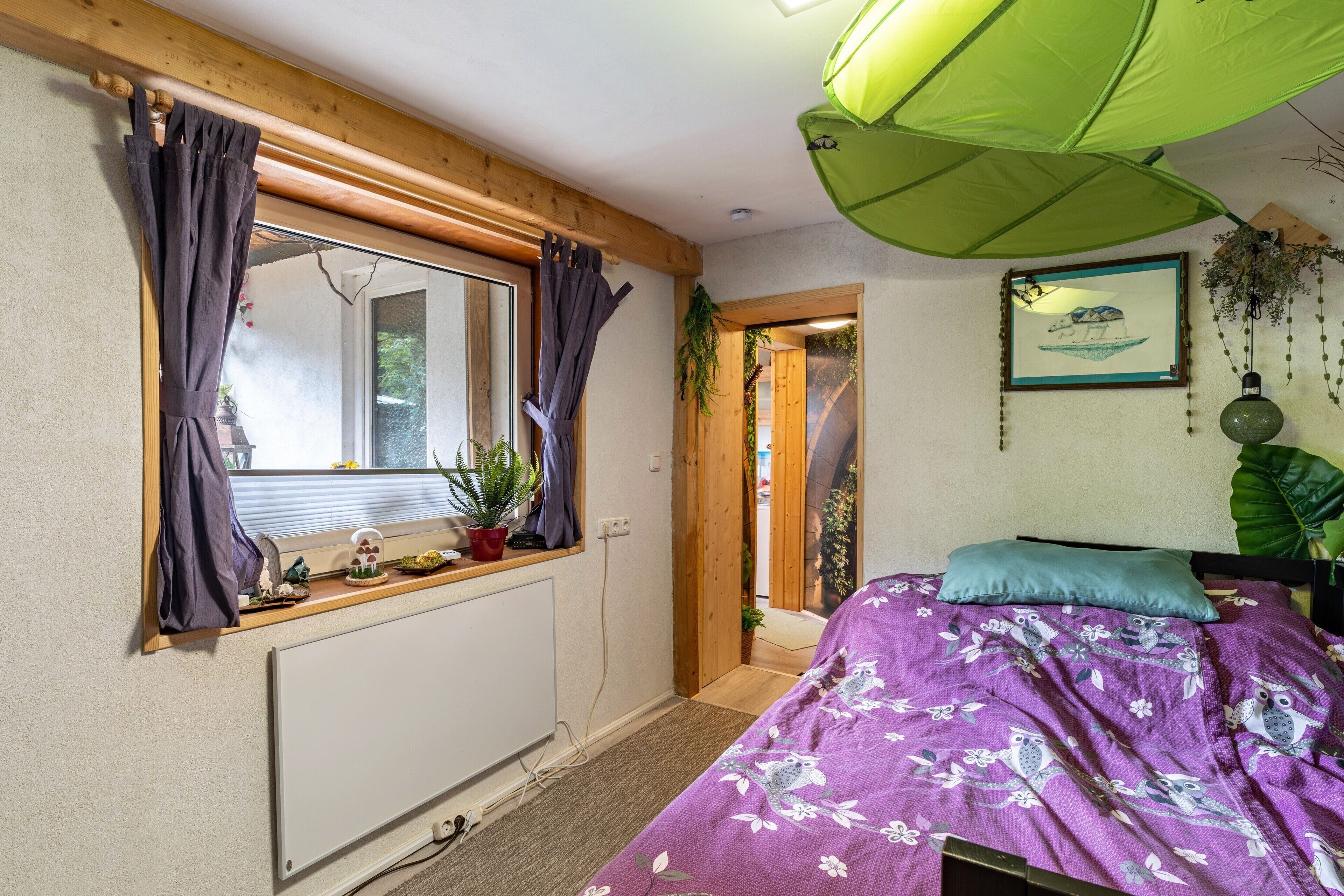 A bedroom with a bed, a window with curtains, a radiator, a green umbrella, and a door leading to another room.